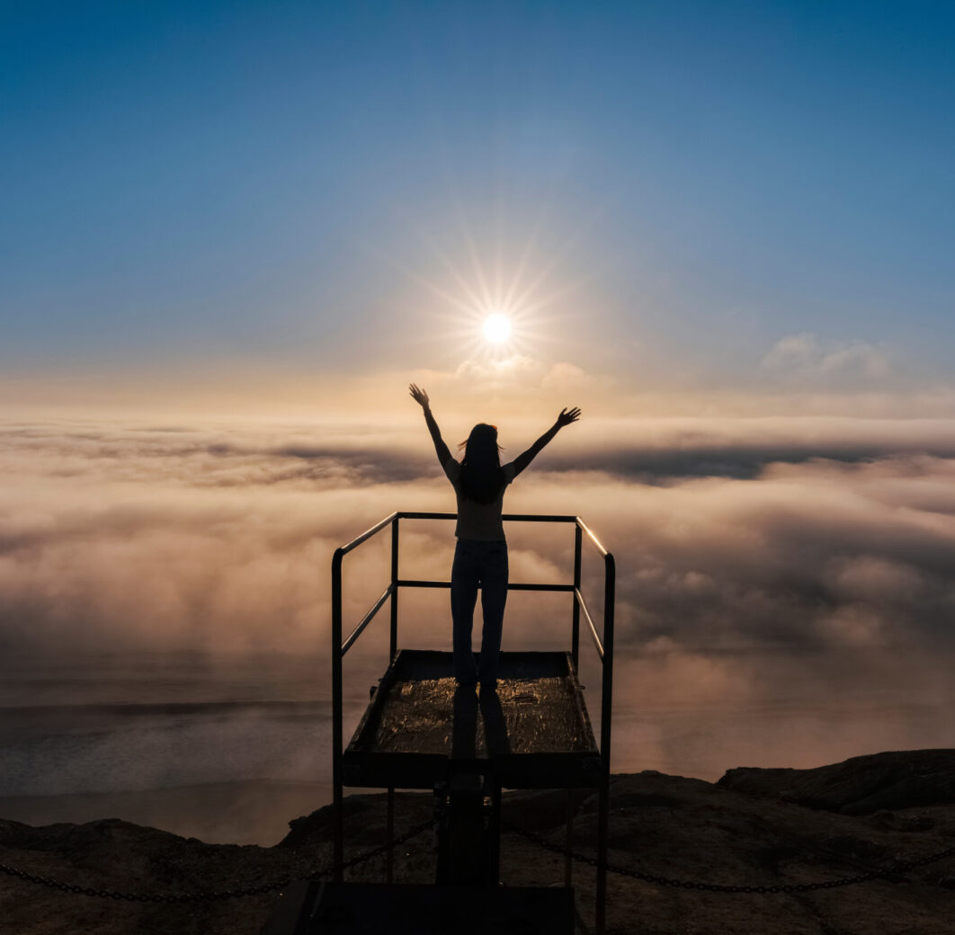 Woman stands on an extended platform high above the clouds with arms outstretched to the sky