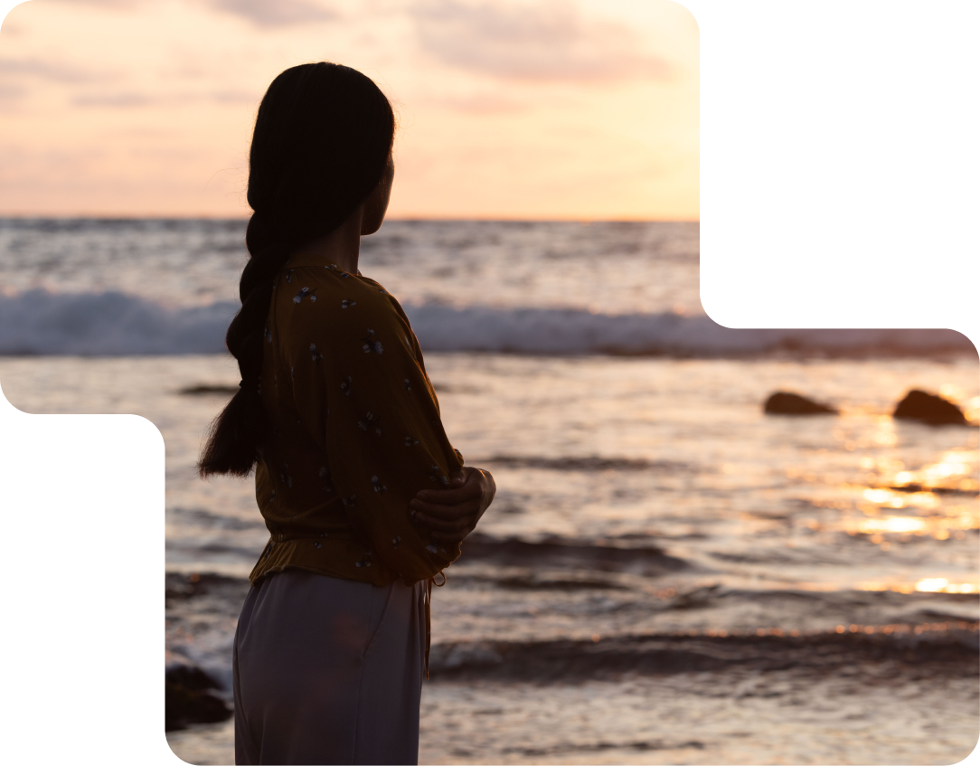 Woman shrouded in shadow on a beach stares out at the ocean at sunset