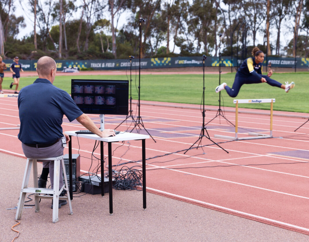 A man sits in front of a computer monitor at the edge of a running track as he analyzes the performance of the hurdle jumper running past him