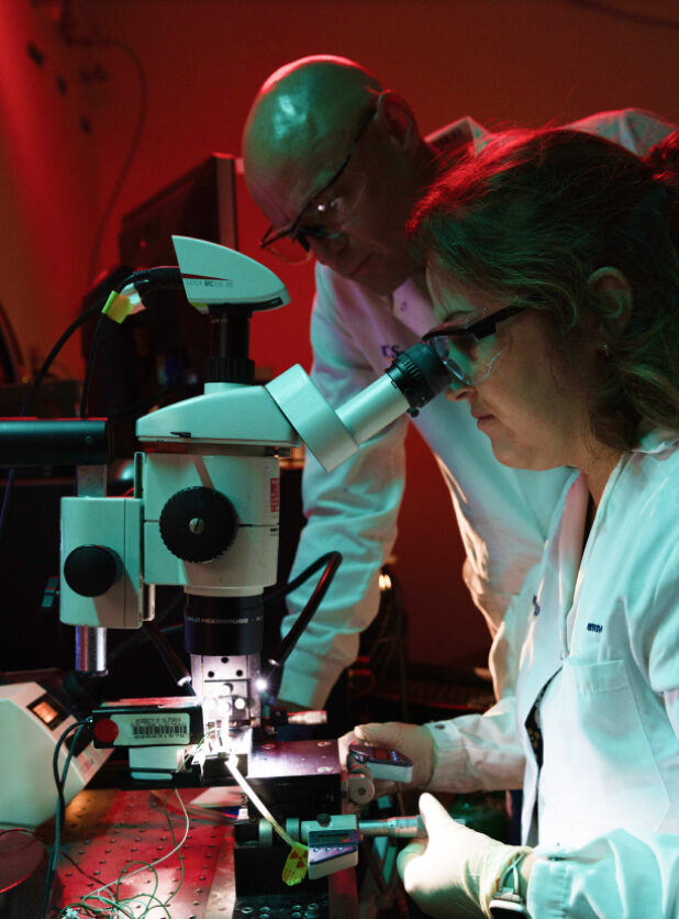 Two scientists in a darkened lab review a specimen using a microscope