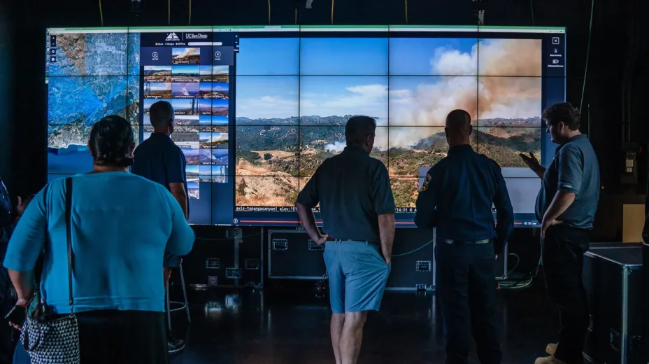 A group of people stand in front of a wall of screens showing a potential wildfire in the wilderness, along with views from other cameras displayed on smaller screens